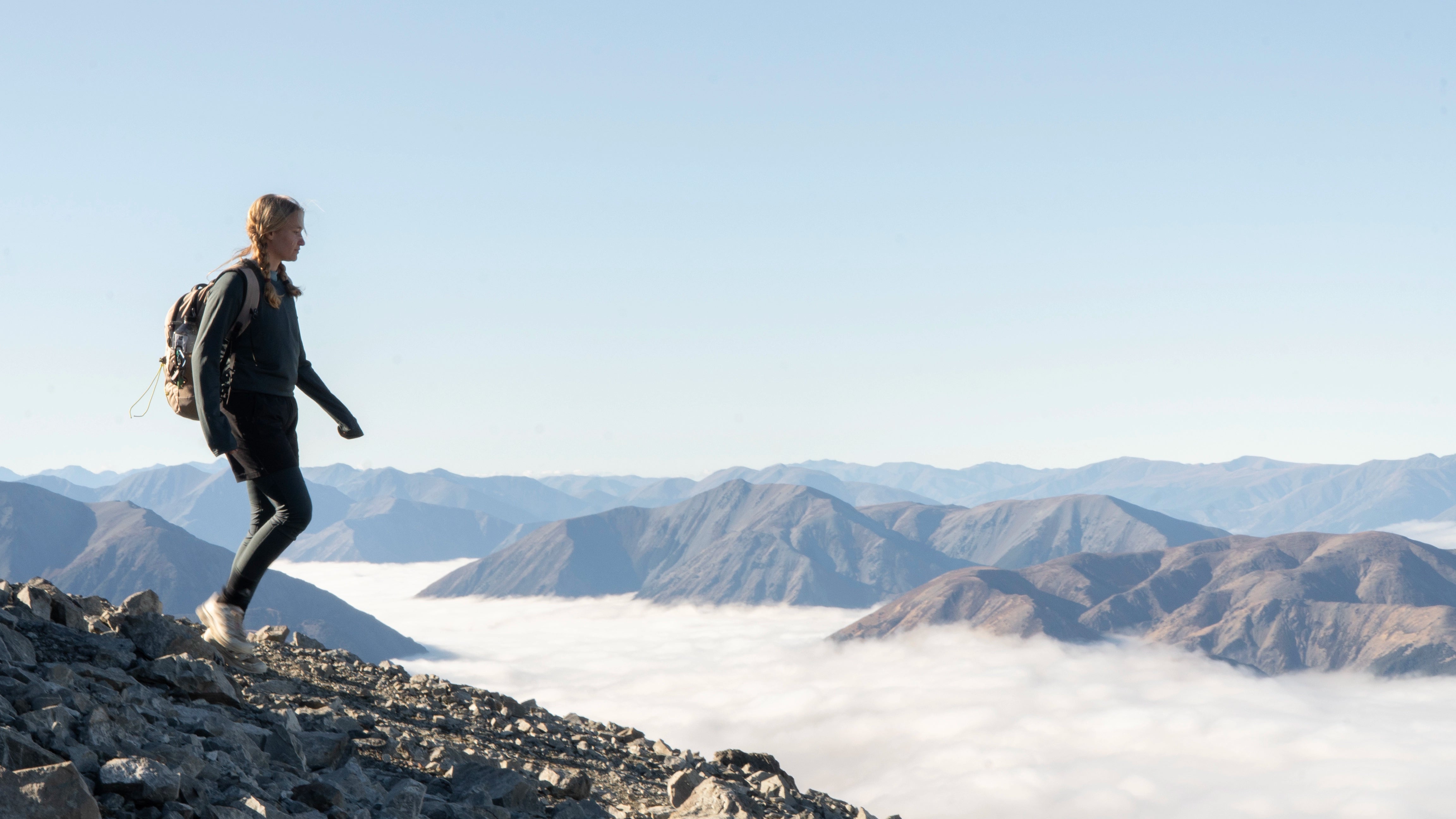 Person hiking on a mountain with a backpack, overlooking a scenic landscape with mountains and clouds.
